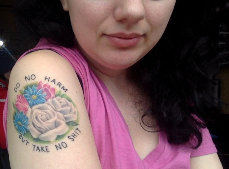 Kate in a pink shirt, showing off an upper arm tattoo that says "Do No Harm, But Take No Shit" and has pink tulips, blue daisies, and white roses